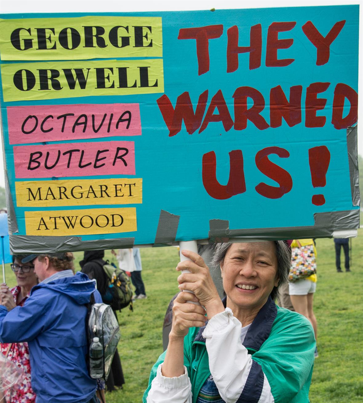Picture of person in dispersed crowd holding picket sign that reads George Orwell Octavia Butler Margaret Atwood They Warned Us
